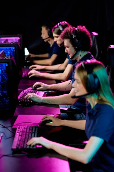 A row of players in noise-cancelling headsets, seated side by side and heads-down on their games under pink and blue light